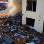 Casa danificada por explosão de botijão de gás em Ceilândia, com fumaça, destroços e viatura de bombeiros ao fundo.