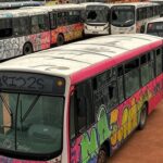 Ônibus vandalizados em pátio no Distrito Federal, com danos como vidros quebrados e pichações, em estilo fotojornalístico.