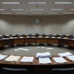 Sala de sessões vazia na CLDF com pilhas de documentos e relógio parado, representando lentidão burocrática e falta de avanços.