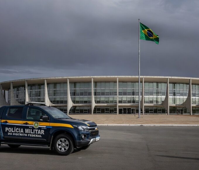Edifício da CLDF em Brasília com símbolos de combate à violência contra a mulher e viatura policial, destacando falhas na reintegração de policiais.