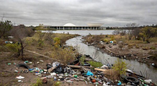 Área ambiental degradada no DF com lixo e vegetação poluída, ao fundo prédio da CLDF, destacando falhas persistentes em meio a homenagens.