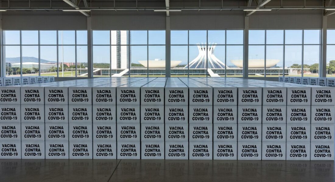 Caixas de vacinas contra Covid-19 em armazém no Distrito Federal, com arquitetura de Brasília ao fundo.