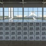 Caixas de vacinas contra Covid-19 em armazém no Distrito Federal, com arquitetura de Brasília ao fundo.