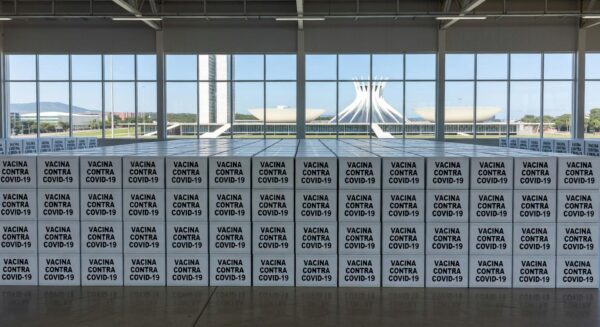 Caixas de vacinas contra Covid-19 em armazém no Distrito Federal, com arquitetura de Brasília ao fundo.
